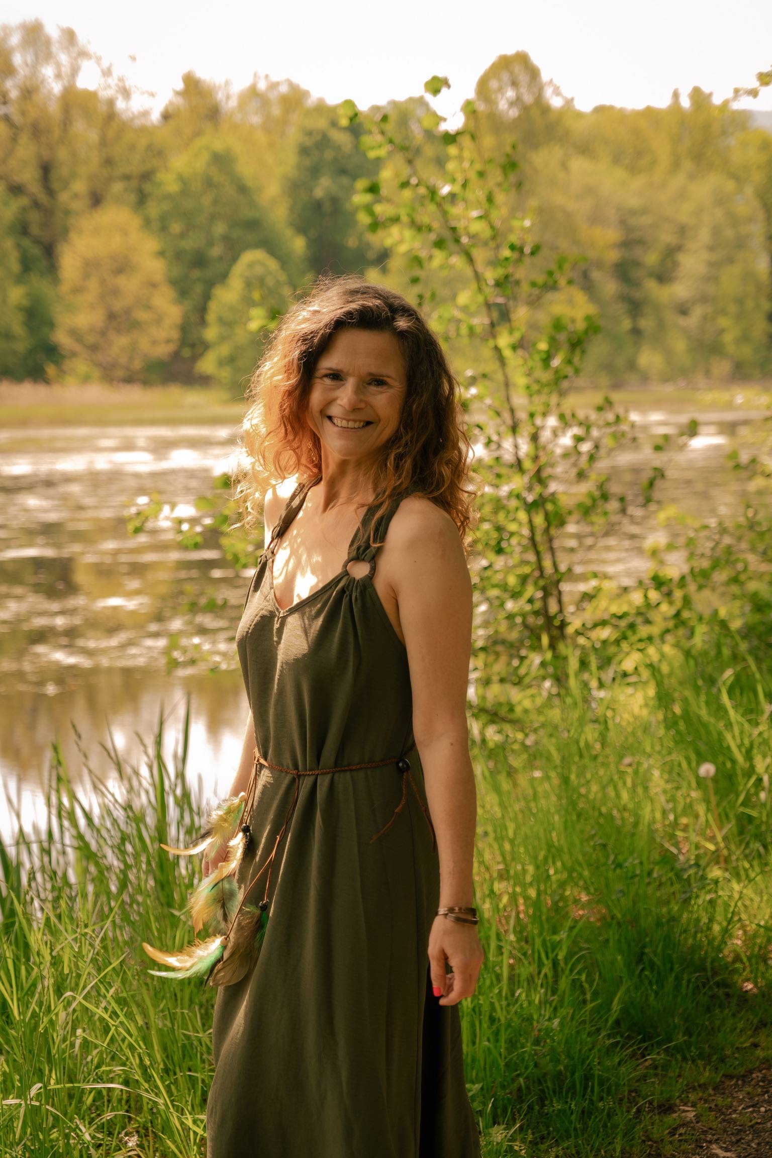 Anna Kuśmirek w oliwkowej sukni nad stawem, w złotym świetle pod słońce, patrzy w obiektyw i uśmiecha się