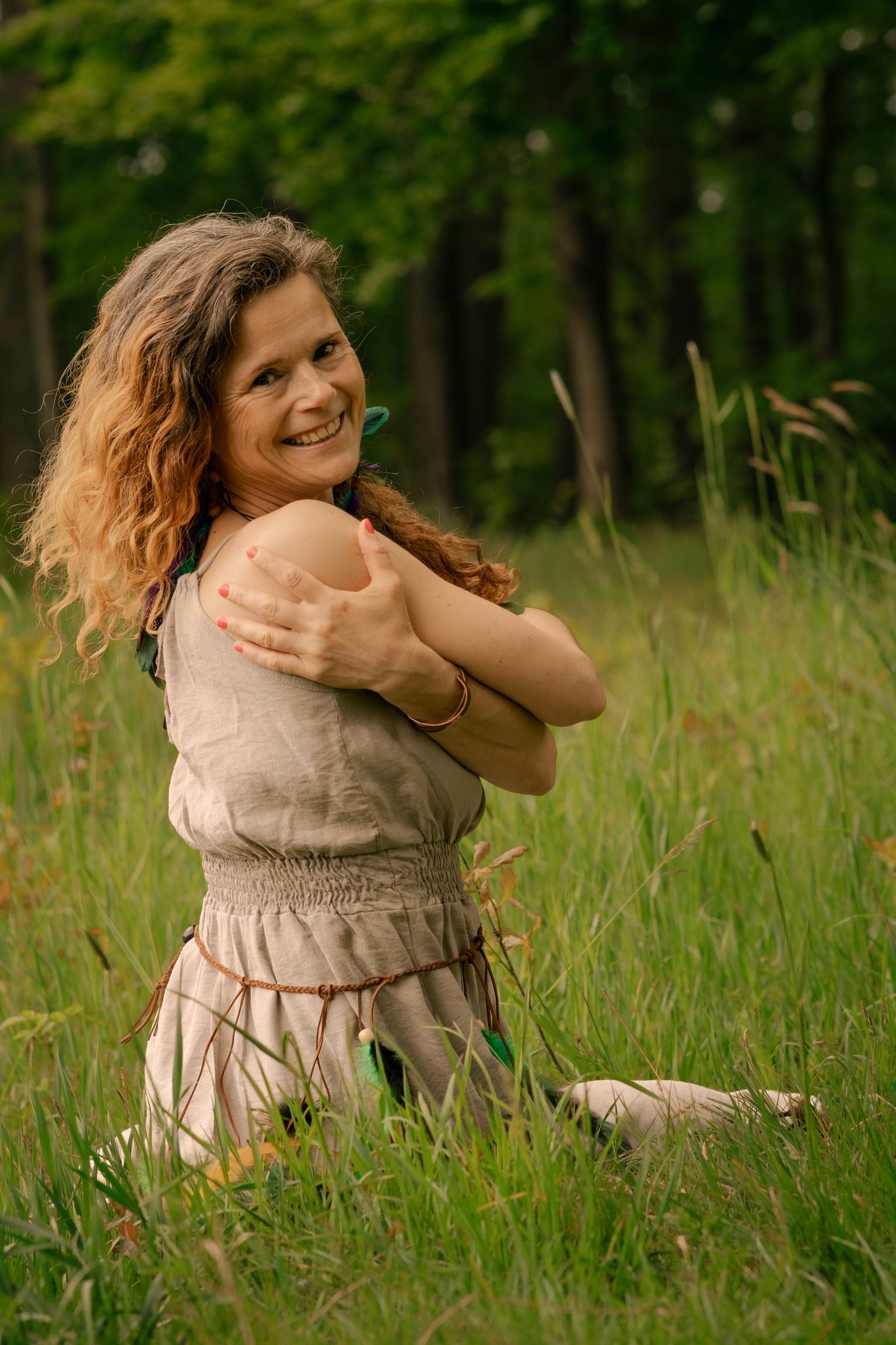 Anna Kuśmirek w lnianej sukience, z piórami we włosach, obejmuje ramiona i uśmiecha się przez ramię w wysokiej trawie